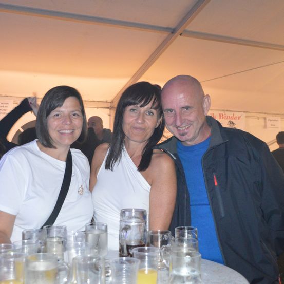 Drei Personen, zwei Frauen und ein Mann, lächeln und posieren für ein Foto in einem Zelt mit verschwommenem Hintergrund. Sie sind von Gläsern mit Getränken umgeben.