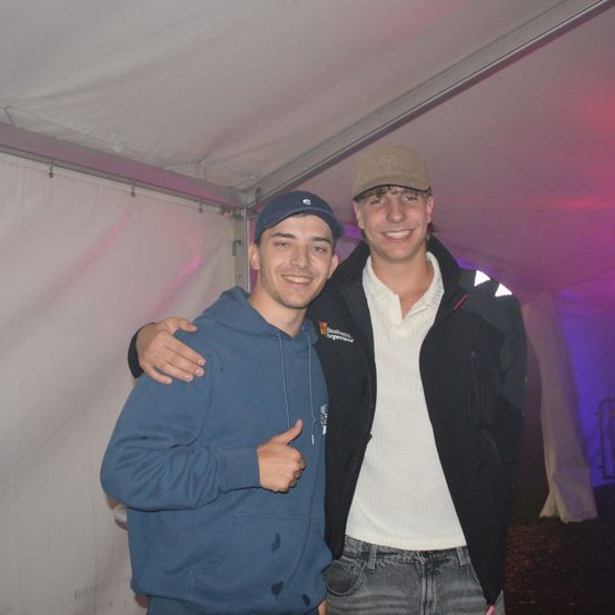 Zwei Männer stehen in einem Zelt, einer zeigt den Daumen hoch. Einer trägt eine blaue Kapuzenjacke, der andere eine schwarze Jacke und ein weißes Hemd.