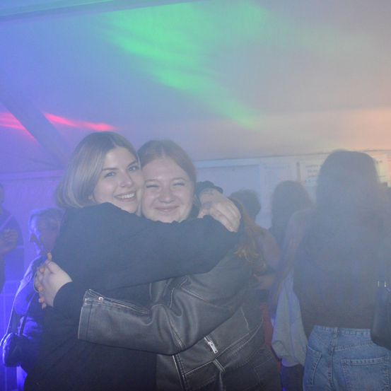 Zwei Frauen umarmen sich auf einer Party, lächeln und posieren für ein Foto. Der Hintergrund ist schwach beleuchtet mit farbigen Lichtern.