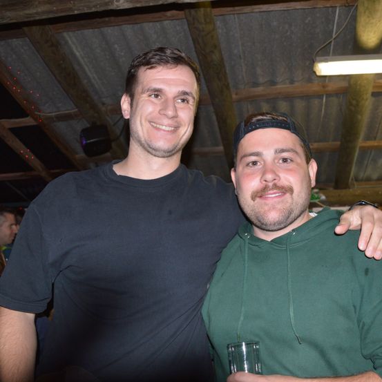 Zwei Männer, die für ein Foto lächeln, einer in einem schwarzen T-Shirt und der andere in einer grünen Kapuzenjacke, stehen in einem schwach beleuchteten Raum mit Holzbalken und roten Lichtern.