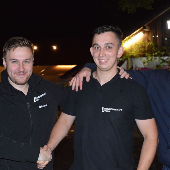 Drei Männer stehen zusammen und lächeln für ein Foto bei Nacht. Der Mann in der Mitte trägt ein schwarzes Poloshirt mit weißem Text. Der Mann auf der linken Seite trägt eine schwarze Jacke. Dahinter befindet sich ein Gebäude mit einem beleuchteten Bereich.