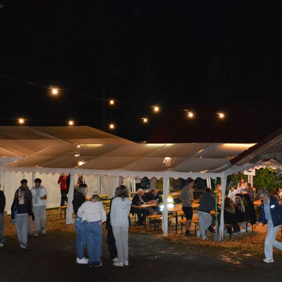 Eine nächtliche Zusammenkunft unter Zelten mit Lichterketten. Einige Personen sitzen an Tischen, andere stehen. Der Bereich ist gut beleuchtet.