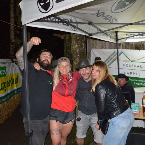 Vier Menschen stehen bei Nacht unter einem Zelt, zwei Frauen und zwei Männer, lächeln für die Kamera. Der Mann auf der linken Seite hat eine Tätowierung am Arm.