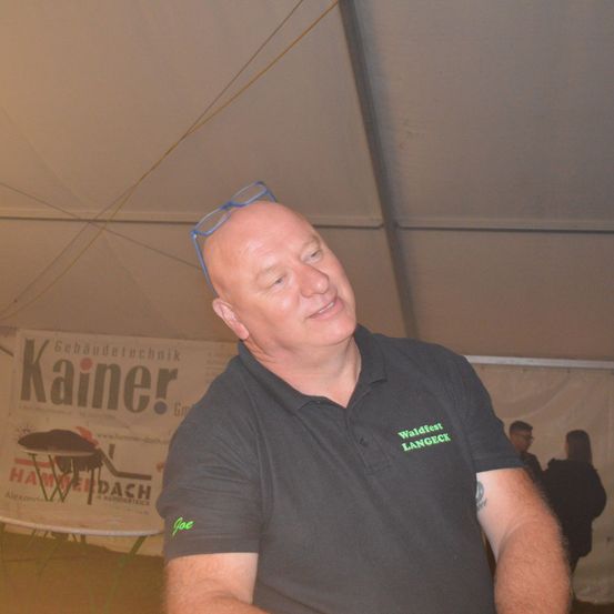 Ein Mann steht unter einem Zelt und trägt ein schwarzes Shirt mit dem Wort Joe und dem Logo Waldfest Langeck. Er hat eine Brille auf dem Kopf. Hinter ihm wird ein Banner mit dem Wort Kainer angezeigt. Hinter dem Banner stehen Menschen.