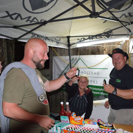 Drei Männer unter einem Zelt, zwei halten kleine Flaschen und lächeln. Hinter ihnen steht ein Banner mit Holzbau Pel. Verschiedene Gegenstände, einschließlich Ketchupflaschen, sind auf dem Tisch.