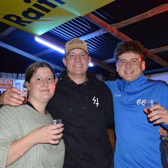 Drei Personen posieren für ein Foto in einem Raum mit einem gelben Banner, das 'Rain' sagt. Zwei Männer lächeln und die Frau hält ein Glas. Der Mann in der Mitte trägt eine Baseballkappe.