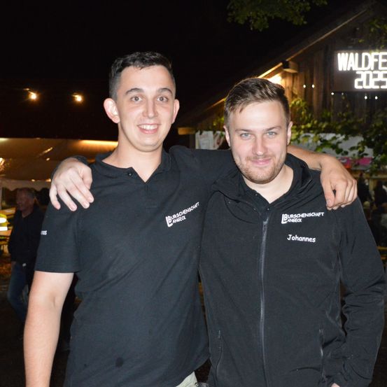Zwei Männer stehen nachts draußen, einer trägt ein schwarzes Poloshirt mit weißem Text, der andere ein schwarzes Jacket mit dem Namen Johannes. Hinter ihnen hat ein Gebäude ein Schild mit dem Text Waldfest 2023.