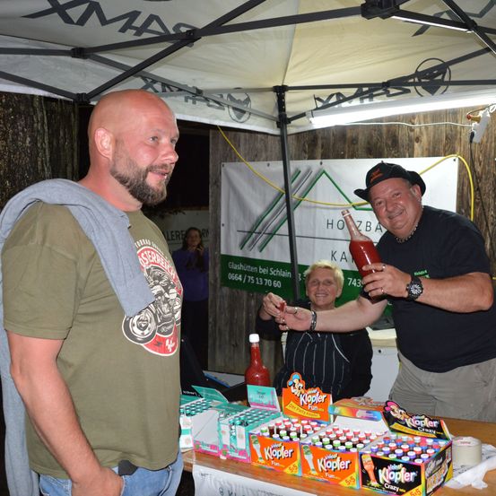 Ein Mann in einem grünen T-Shirt lächelt und unterhält sich mit einem anderen Mann, der eine Flasche Ketchup hält. Hinter ihnen steht eine Frau, die eine Flasche hält. Sie stehen unter einem Zelt.