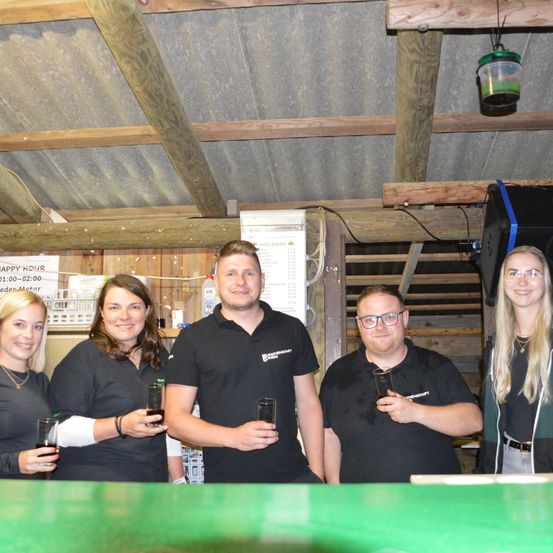 Fünf Personen stehen zusammen an einer Bar, lächeln und halten Gläser. Sie tragen schwarze Shirts, außer eine mit einer grünen Jacke. Ein Schild an der Wand steht für Happy Hour.