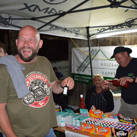 Ein Mann in einem grünen T-Shirt mit einem Motorraddesign hält einen Schlüssel. Neben ihm gießt ein anderer Mann eine Flasche Ketchup ein. Eine ältere Frau steht hinter einem Tisch voller Produktboxen. Sie stehen unter einem Zelt mit einem Banner, das Holzbauer liest.