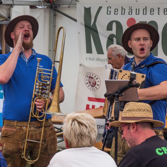 Zwei Männer in blauen Hemden und braunen Hüten spielen Musik mit einer Posaune und einem Akkordeon. Ein älterer Mann steht hinter ihnen, und ein Mann mit einem Strohhut hört zu. Ein Banner mit deutschem Text ist im Hintergrund.