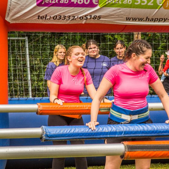 Eine Gruppe von Frauen in pinken T-Shirts nimmt an einem aufblasbaren Spiel im Freien teil. Ein Banner mit Preisen und Kontaktinformationen hängt oben.