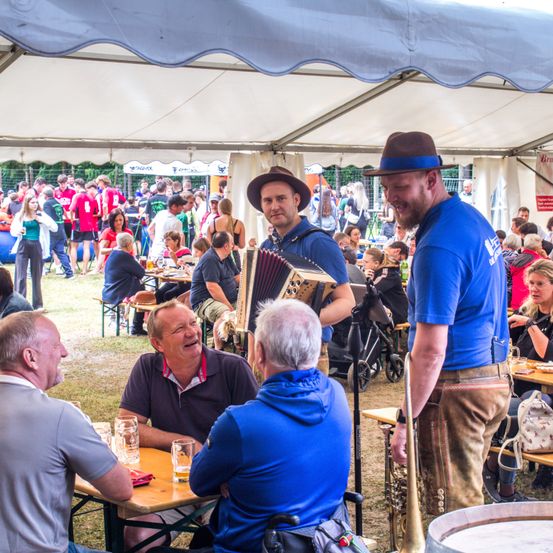 Eine Gruppe von Menschen genießt ein Outdoor-Festival. Drei Männer in Blau unterhalten sich in der Nähe eines Tisches mit Biergläsern. Zwei Männer mit Akkordeons und einer Trompete spielen. Viele Menschen sitzen und stehen und beobachten die Aufführung.