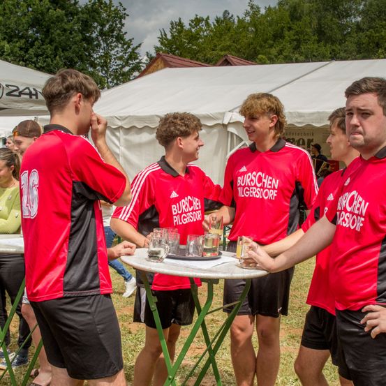 Eine Gruppe von Menschen in roten T-Shirts mit weißem Text steht um einen Tisch mit Gläsern. Sie befinden sich bei einer Outdoor-Veranstaltung.