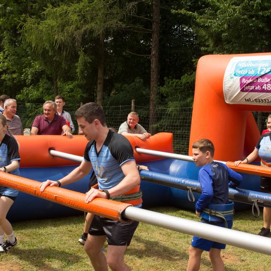 Eine Gruppe von Menschen nimmt an einem aufblasbaren Hindernisparcours im Freien teil, während Zuschauer zusehen und anfeuern.