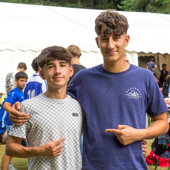 Zwei Jungen posieren für ein Foto bei einer Outdoor-Veranstaltung. Ein Junge trägt ein blaues Shirt mit Logo, der andere ein kariertes. Menschen sind im Hintergrund.