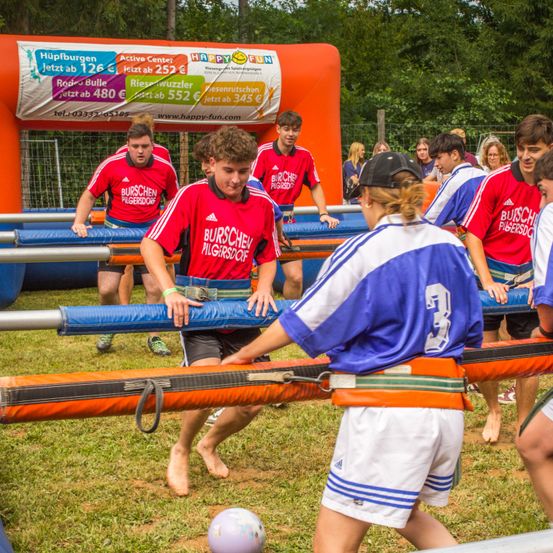 Eine Gruppe junger Männer und Frauen spielt draußen auf einem Rasenfeld ein aufblasbares Dodgeball-Spiel. Das Spiel wird von Happy Fun gesponsert.