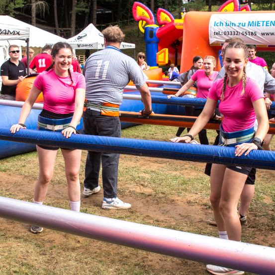 Eine Gruppe von Menschen nimmt an einer Outdoor-Veranstaltung teil. Zwei Frauen stehen und halten sich an einem blauen Rohr fest, während sie lächeln. Ein erwachsener Mann steht hinter ihnen und beobachtet. Im Hintergrund befinden sich aufblasbare Hindernisse und Zelte.