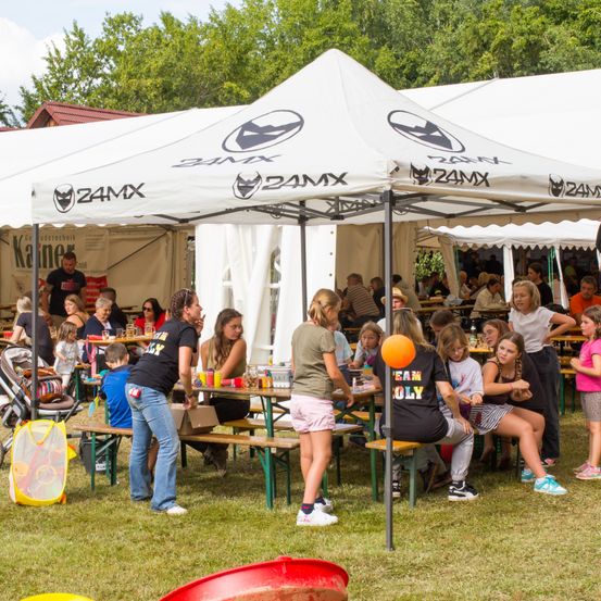 Eine Gruppe von Menschen isst an einem Tisch unter einem Zelt mit der Aufschrift 24MX. Einige sitzen, andere stehen. Eine Person hält einen orangefarbenen Luftballon. Das Zelt hat weiße Wände und ein rotes Dach.