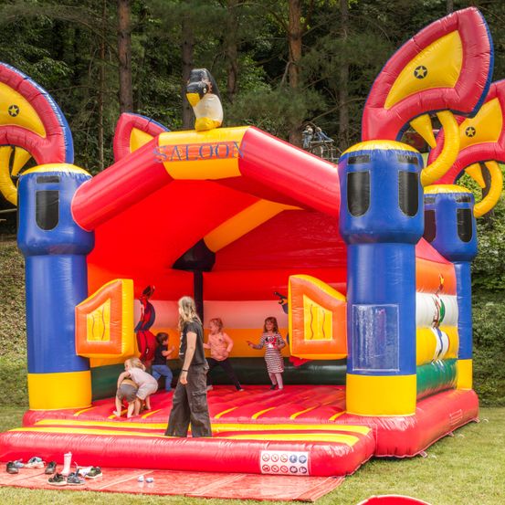 Eine Gruppe von Kindern spielt in einem großen aufblasbaren Spielhaus mit der Aufschrift Saloon. Eine Frau steht in der Nähe und beobachtet sie. Das Spielhaus ist leuchtend in Rot, Blau und Gelb gehalten. Vor dem Spielhaus liegen Schuhe auf dem Gras verstreut.