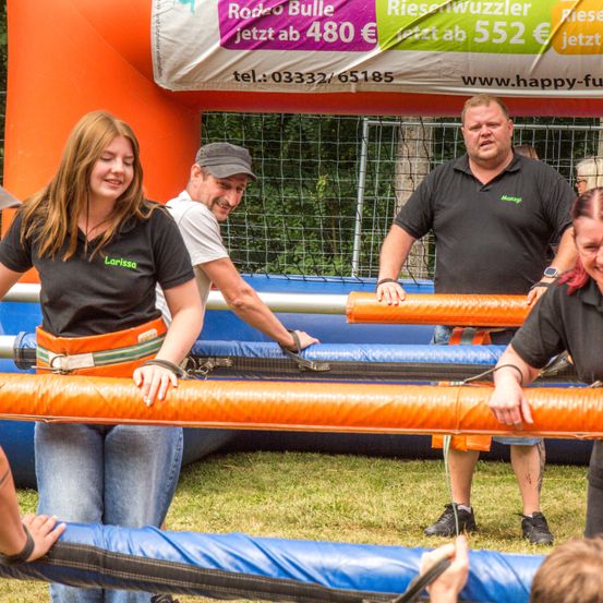 Mehrere Erwachsene befinden sich bei einer karnevalähnlichen Veranstaltung, zwei Männer reparieren etwas, während eine Frau vor ihnen steht. Sie sind in der Nähe eines Sprunghauses und ein Schild mit Preisen steht hinter ihnen.