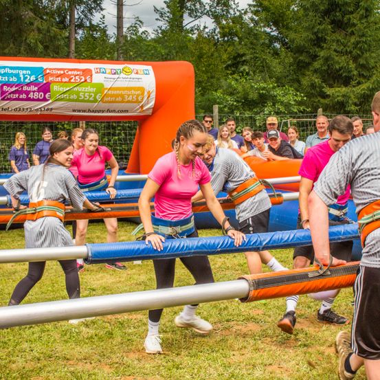 Mehrere Personen spielen ein aufblasbares Spiel auf einem Rasenfeld. Sie halten sich an einem langen blauen Stab. Sie tragen Gürtel und Turnschuhe. Dahinter befindet sich eine große orangefarbene aufblasbare Struktur.
