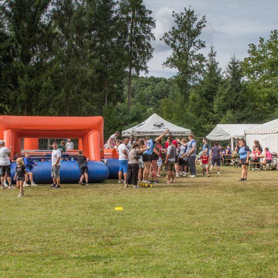 Eine Gruppe von Menschen steht und versammelt sich um aufblasbare und mit Netzen versehene Spiele auf einer Rasenfläche, mit mehreren Zelten im Hintergrund.