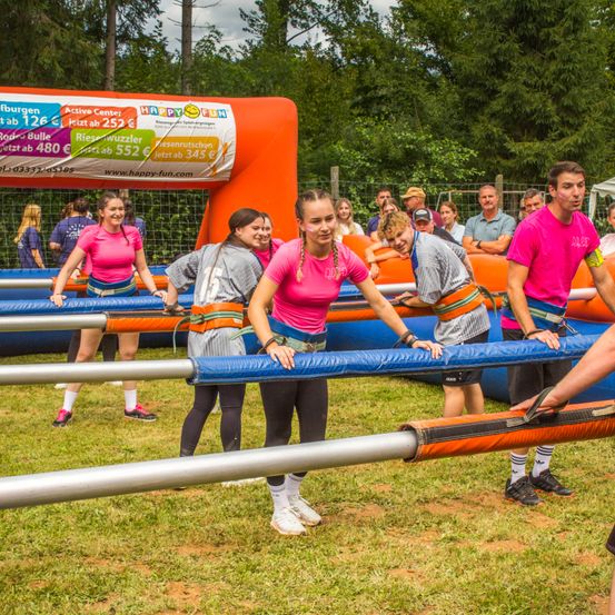 Eine Gruppe von Menschen nimmt an einer Outdoor-Hindernisparcours-Veranstaltung teil. Mehrere Personen halten große Stangen und tragen pinke Shirts. Dahinter befindet sich ein orangefarbener aufblasbarer Hindernisparcours mit Werbetext.