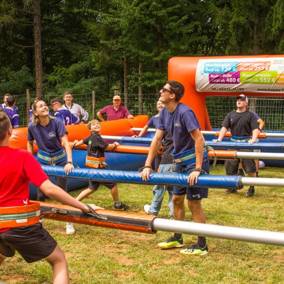 Mehrere Personen in blauen und roten Uniformen nehmen an einer Teambuilding-Aktivität im Freien teil, mit aufblasbaren Hindernissen und Bäumen im Hintergrund.