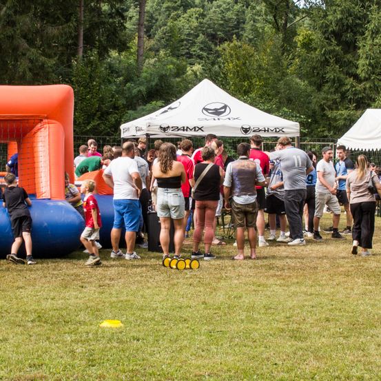 Eine Gruppe von Menschen versammelt sich im Freien, einige stehen und andere sitzen. Links ist ein aufblasbarer Hindernisparcours aufgebaut. Es gibt Zelte mit Markenzeichen, und die Menschen sind in verschiedenen Aktivitäten verstrickt.