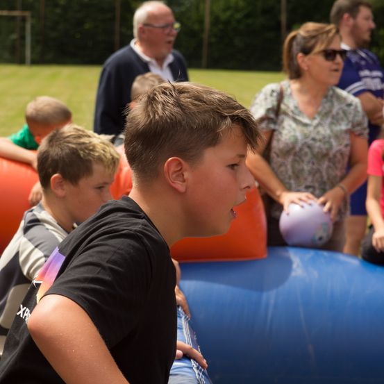 Eine Gruppe von Kindern und Erwachsenen steht neben aufblasbaren Hindernissen. Ein Junge in Schwarz schaut nach vorne, während ein anderer einen lila Ball hält.