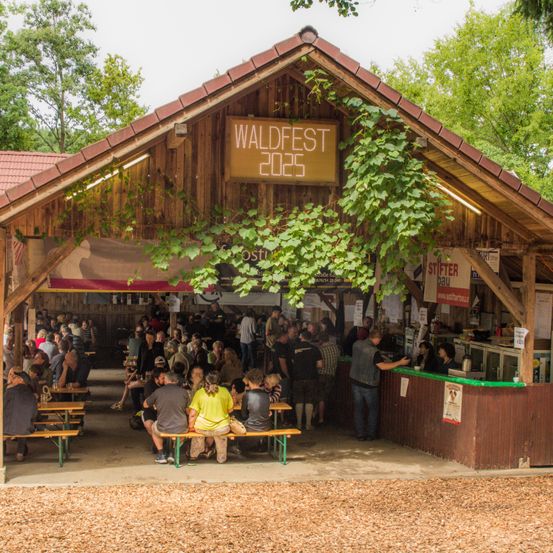 Eine Gruppe von Menschen hat sich in einem hölzernen Pavillon versammelt. Der Pavillon trägt ein Schild mit der Aufschrift 'WALDFEST 2023'. Die Leute sitzen auf Bänken und stehen herum, wahrscheinlich auf etwas wartend. Es gibt einen Tresen mit einer Frau dahinter.