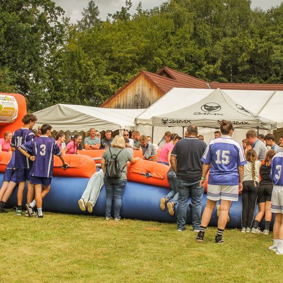 Eine Gruppe von Menschen steht auf einer Rasenfläche, einige tragen Trikots mit Nummern. In der Nähe sind aufblasbare Hindernisse aufgestellt. Einige Personen sitzen auf den aufblasbaren Geräten. Im Hintergrund sind mehrere Zelte zu sehen.