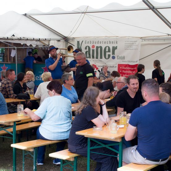 Eine Gruppe von Menschen sitzt an Tischen bei einer Outdoor-Veranstaltung unter einem Zelt. Ein Mann in einem blauen Shirt spielt Trompete, während andere ihre Getränke genießen.