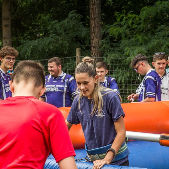 Eine Gruppe von Menschen, einige in blauen Trikots und andere in Freizeitkleidung, versammelt sich um ein aufblasbares Hindernis. Eine Frau in einem blauen Shirt schaut einen Mann in einem roten Shirt an.
