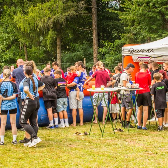 Eine Gruppe von Menschen steht um einen aufblasbaren Hindernisparcours mit einem Baldachin, Getränken und Tischen. Bäume und ein Zaun sind im Hintergrund.