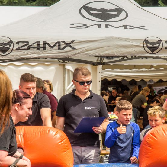 Eine Gruppe von Menschen unter einem Zelt mit dem Logo 24MX. Ein Mann mit Sonnenbrille hält eine Mappe und spricht mit einem Jungen, der einen Daumen hochgibt.