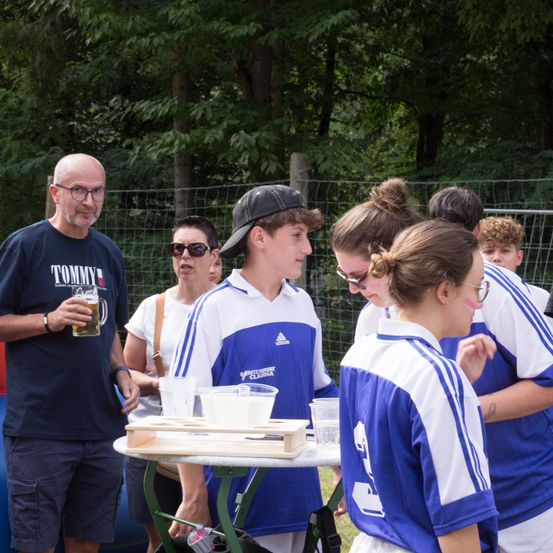 Eine Gruppe von Menschen steht um einen Tisch mit Bechern und Bier. Ein älterer Mann hält ein Glas Bier. Ein Junge und ein Mädchen schauen sich etwas auf dem Tisch an. Ein Zaun und Bäume sind im Hintergrund.