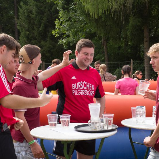 Eine Gruppe junger Männer steht um einen Tisch mit gefüllten Gläsern. Der Mann im roten Shirt mit weißen Texten lächelt und spricht mit anderen. Hinter ihnen befinden sich aufblasbare Objekte.