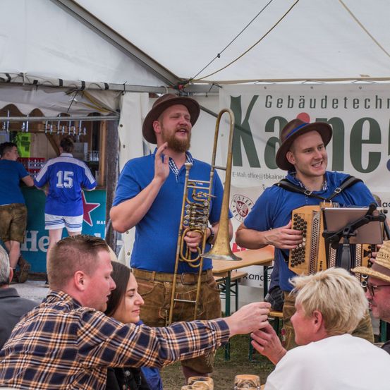 Zwei Männer spielen auf einer Bühne Instrumente, mit einem Publikum, das sich um sie herum versammelt. Einer spielt ein Tenorhorn, und der andere spielt eine Ziehharmonika. Hinter ihnen zeigt ein Banner das Wort 'Kane'.