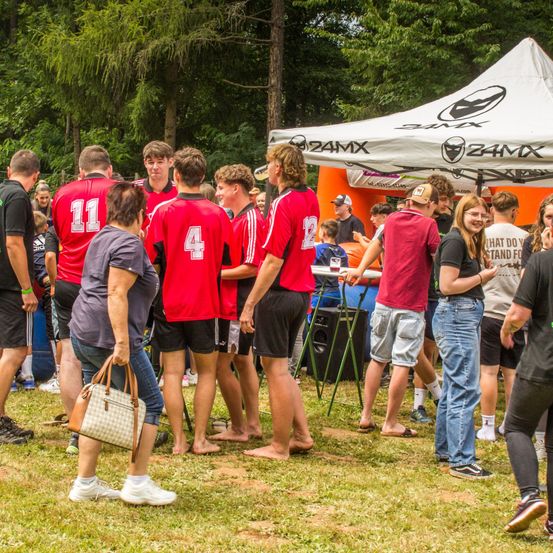 Eine Gruppe junger Leute in roten Trikots steht auf einem Rasen unter einem Zelt und führt wahrscheinlich ein Gespräch. Eine Frau in einem grauen Shirt hält eine Handtasche. In der Nähe spricht ein Mann in einem roten Shirt mit einer Frau mit Brille. Bäume und Menschen sind im Hintergrund zu sehen.