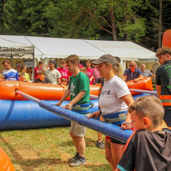 Eine Gruppe von Menschen steht in der Nähe von aufblasbaren Spielen in einem Außenbereich, wobei sich zwei Personen auf eine Aktivität vorbereiten.