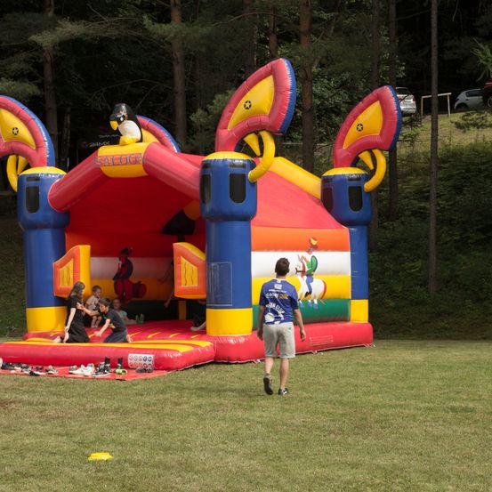 Eine Gruppe von Kindern spielt in einem bunten aufblasbaren Hüpfburg, wobei ein Mann in der Nähe geht. Die Hüpfburg ist mit Zeichentrickfiguren und einem Pinguin-Maskottchen dekoriert.