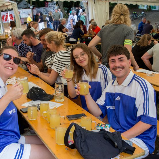 Mehrere Personen in blauen und weißen Uniformen und Gläsern mit orangefarbenen Getränken sitzen an einem Holztisch in einem Biergarten im Freien. Hinter ihnen sitzen weitere Personen an Tischen, und im Hintergrund sind Zelte zu sehen.