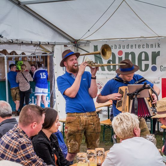 Zwei Männer spielen Musik unter einem Zelt bei einer Veranstaltung im Freien. Einer spielt Trompete, der andere Akkordeon. Zuschauer sitzen um Tische, einige trinken. Im Hintergrund befindet sich eine Bar mit einem Banner.
