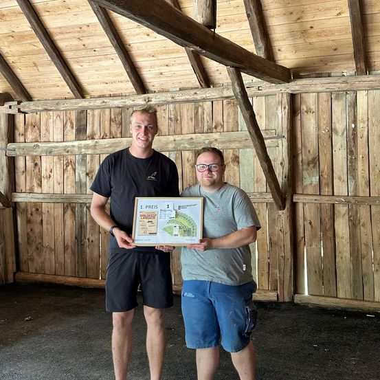 Zwei Männer in einer Scheune halten ein Zertifikat. Einer trägt eine Brille und ein graues T-Shirt, der andere ein schwarzes T-Shirt. Das Zertifikat hat ein Bild und den Text '1. Preis'.