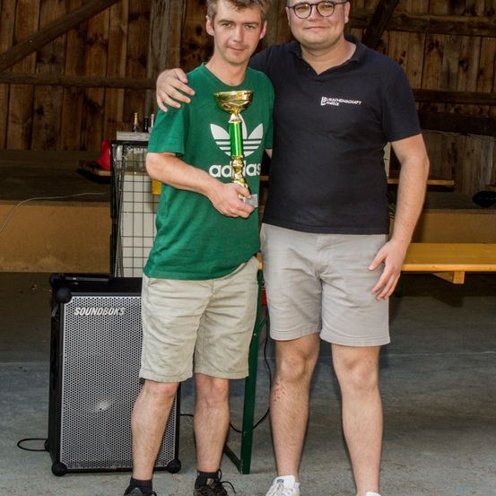 Zwei Männer stehen in einer Scheune; einer hält einen Pokal und der andere steht hinter ihm und lächelt. Der Mann, der den Pokal hält, trägt ein grünes Adidas-Shirt und beige Shorts. Der Mann hinter ihm trägt ein schwarzes Poloshirt mit Logo und khakifarbene Shorts. Ein Lautsprecher steht links und ein Holztisch rechts.