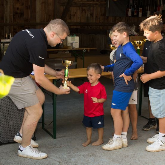 Ein Mann in einem schwarzen Shirt übergibt einem Jungen in einem roten Shirt einen Pokal, während ein Junge in blauen Shorts zusieht. Ein weiterer Junge in einem schwarzen Shirt steht in der Nähe.