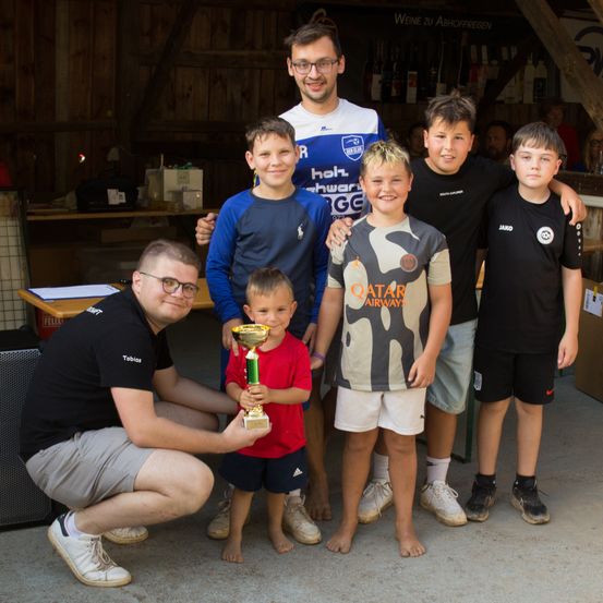 Eine Gruppe von Kindern und Erwachsenen posiert für ein Foto, wobei ein Kind einen Pokal hält. Sie tragen alle Sporttrikots und lächeln.