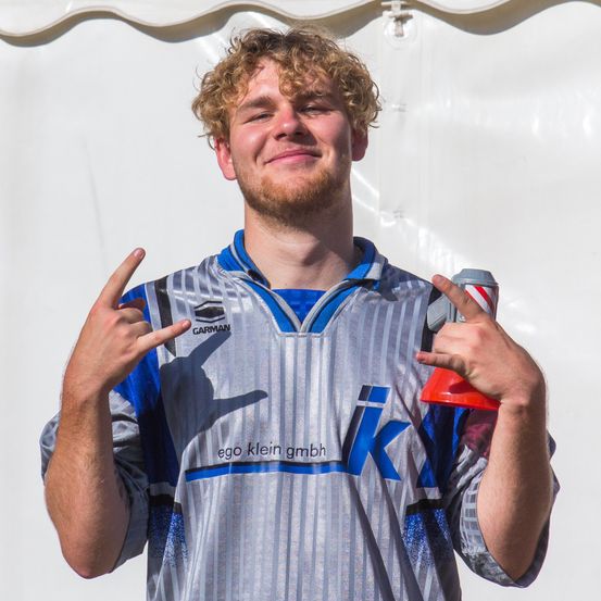 Ein Mann mit lockigem Haar, der ein graues und blaues Sporttrikot trägt, steht und lächelt, hält einen roten und weißen Becher in einer Hand und macht mit der anderen Hand ein Rock-Zeichen.